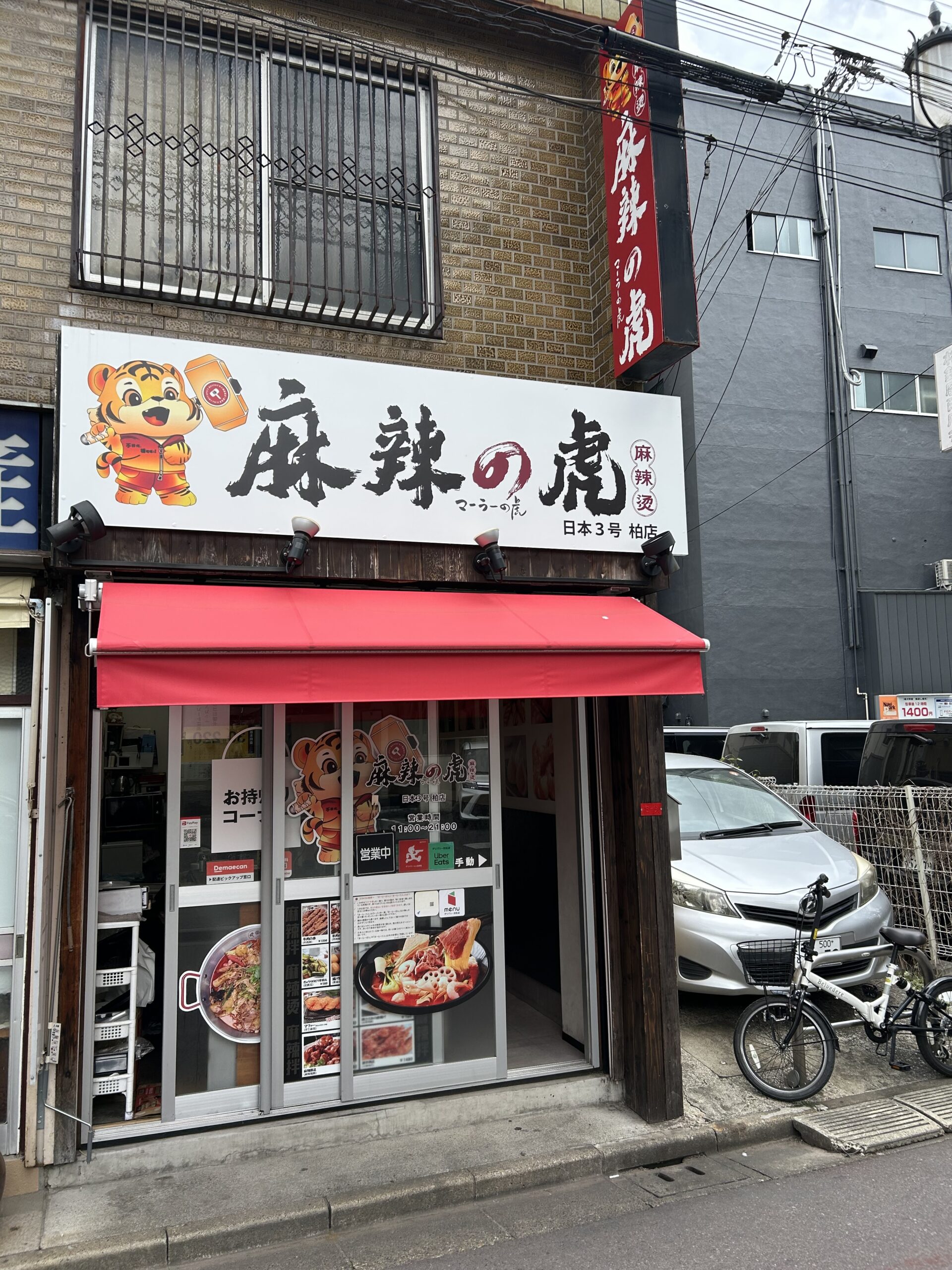 柏・麻辣の虎の店舗外観（白い看板に黒文字で麻辣の虎・トラのキャラクター・赤い暖簾）