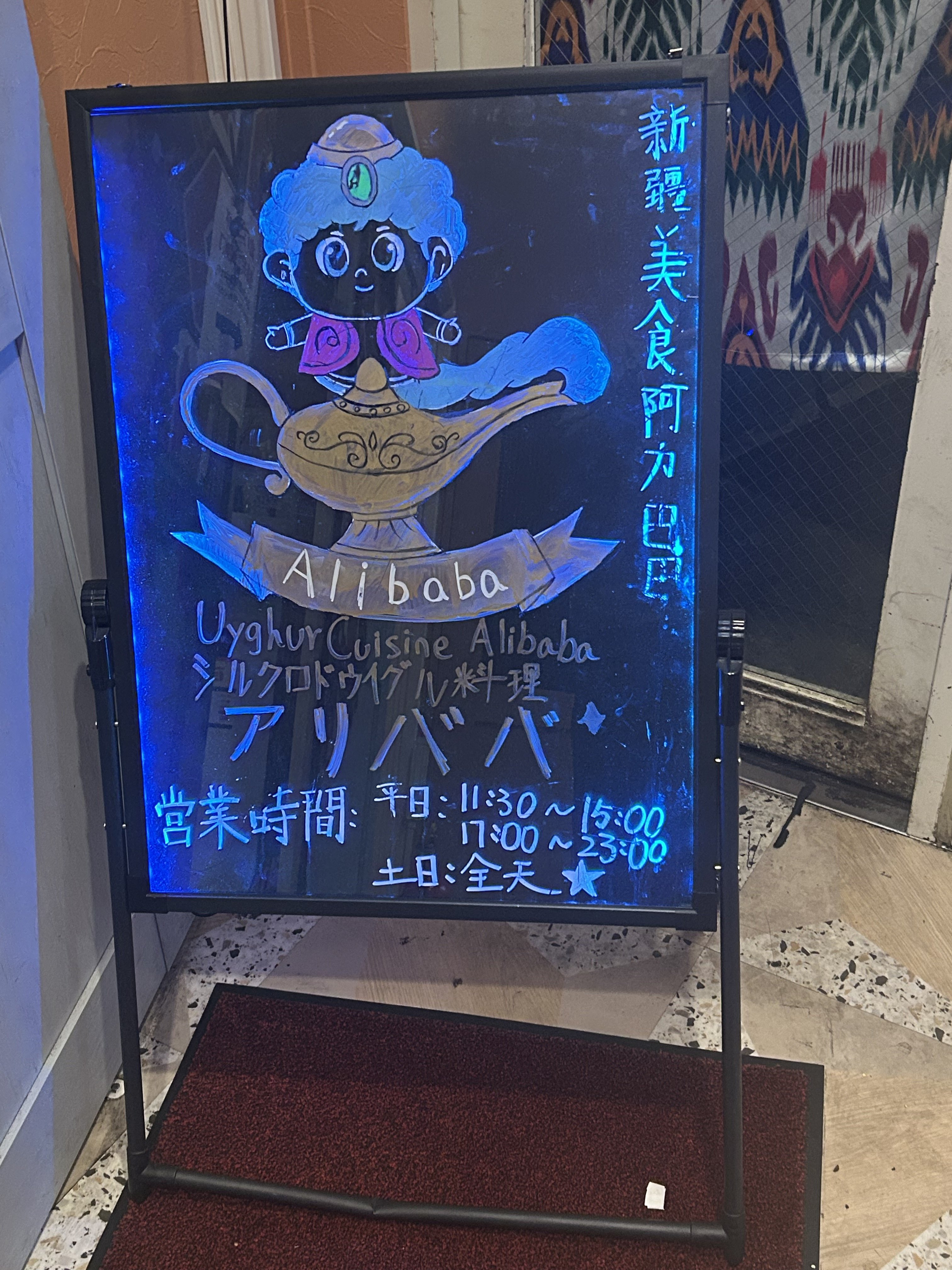 シルクロードウイグル料理 アリババの店頭看板。アラジン風のキャラクターと「新疆美食阿力巴巴」「Uyghur Cuisine Alibaba」「シルクロドウイグル料理 アリババ」の表記